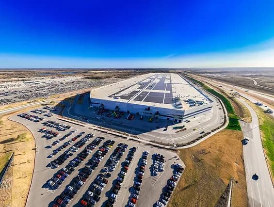 Aerial view of a large warehouse with solar panels, surrounded by a busy parking lot and dry landscape