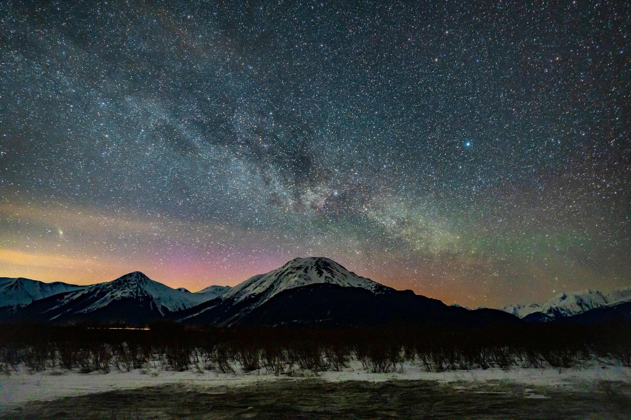 Starry night sky over snow-capped mountains with a dark shrub-filled foreground