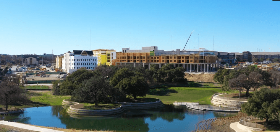 Aerial view of a park with a circular pond and trees, and a construction site with buildings in background