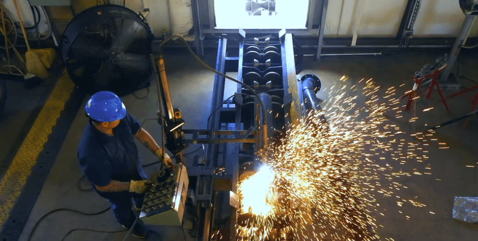 Worker in blue hard hat operates a control panel as sparks fly from a metalworking machine