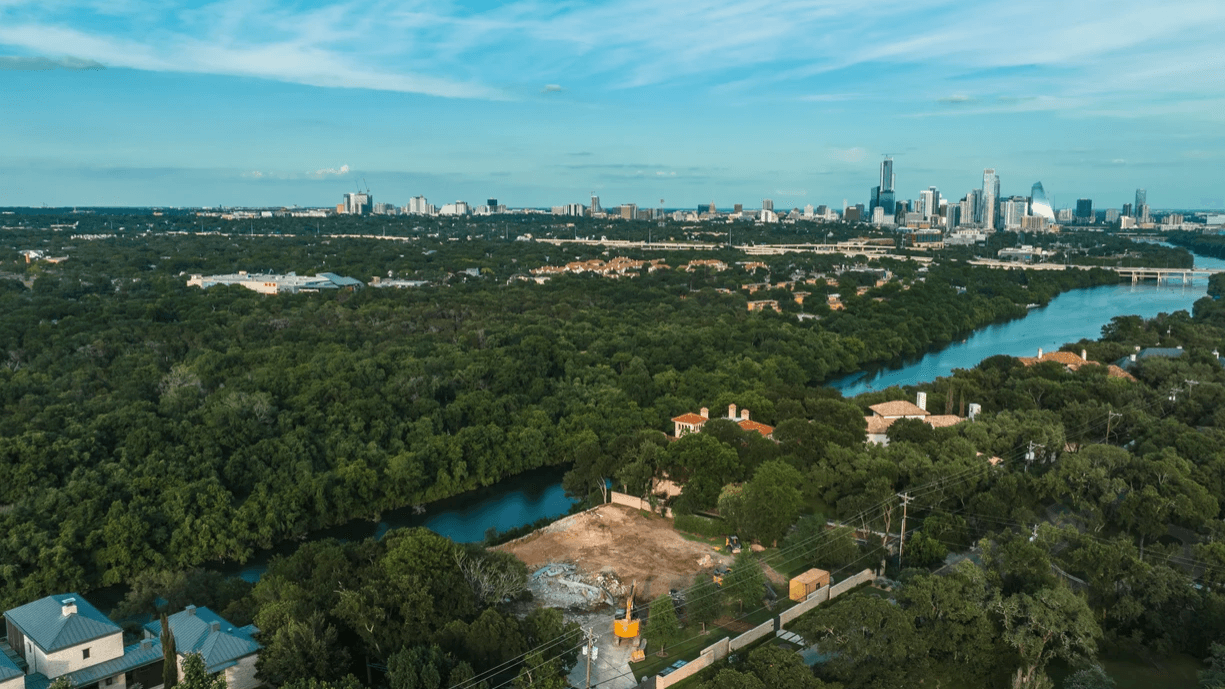 Aerial view of a river winding through a dense forest, with houses and a distant city skyline