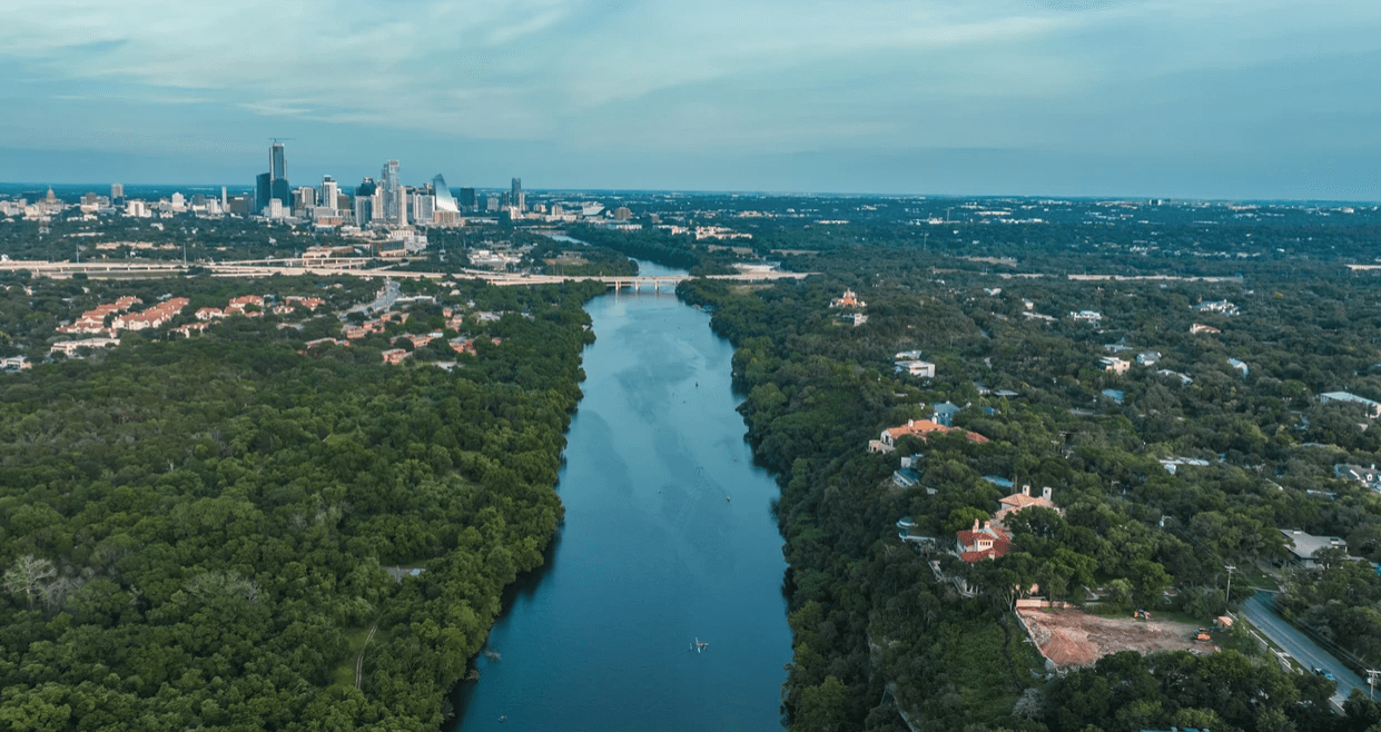 Aerial view of a river winding through dense green forests, with a bridge and city skyline in the distance