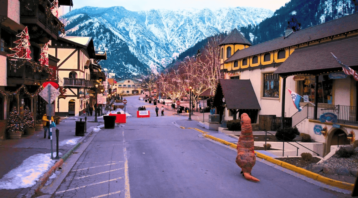 Person in a dinosaur costume on a snow-dusted mountain town street with holiday lights