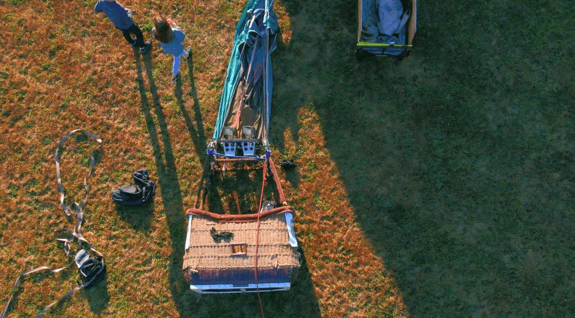 Aerial view of a wicker hot air balloon basket on grass with people and equipment nearby