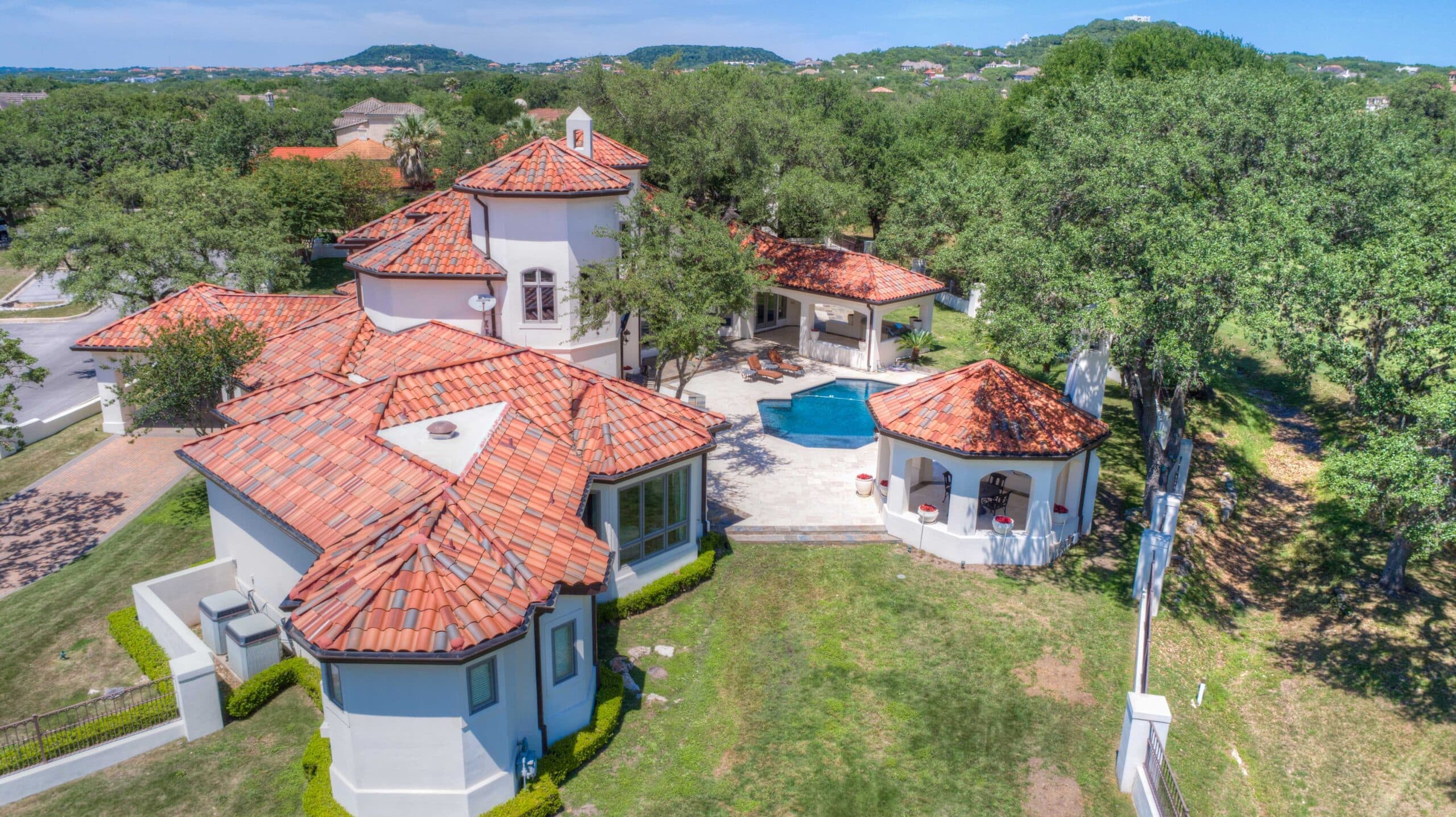 Aerial view of a white stucco villa with red tile roofs, a pool, and patio lounge area amid trees