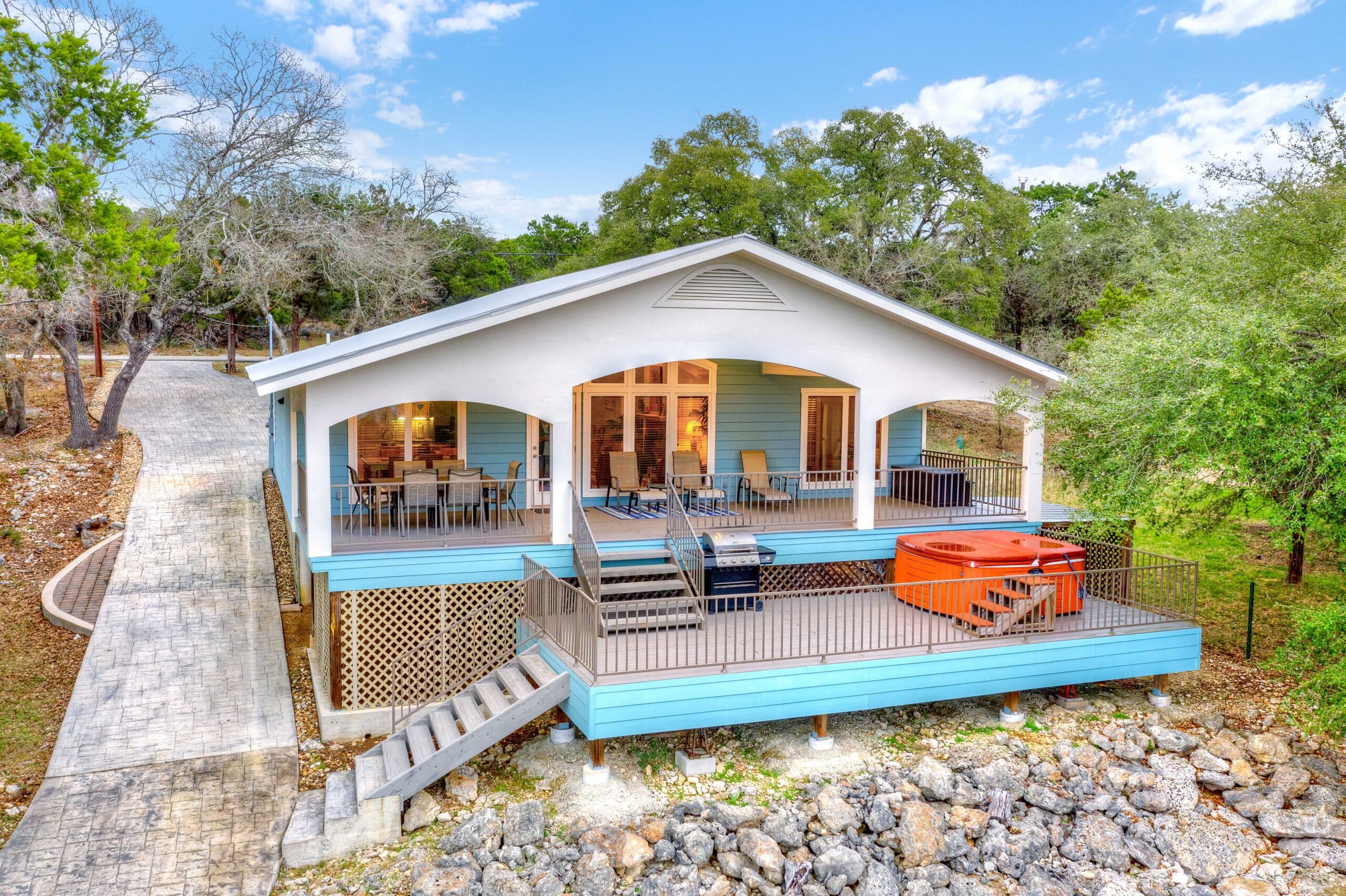 Blue elevated house on stilts with a wraparound porch and outdoor seating, surrounded by trees