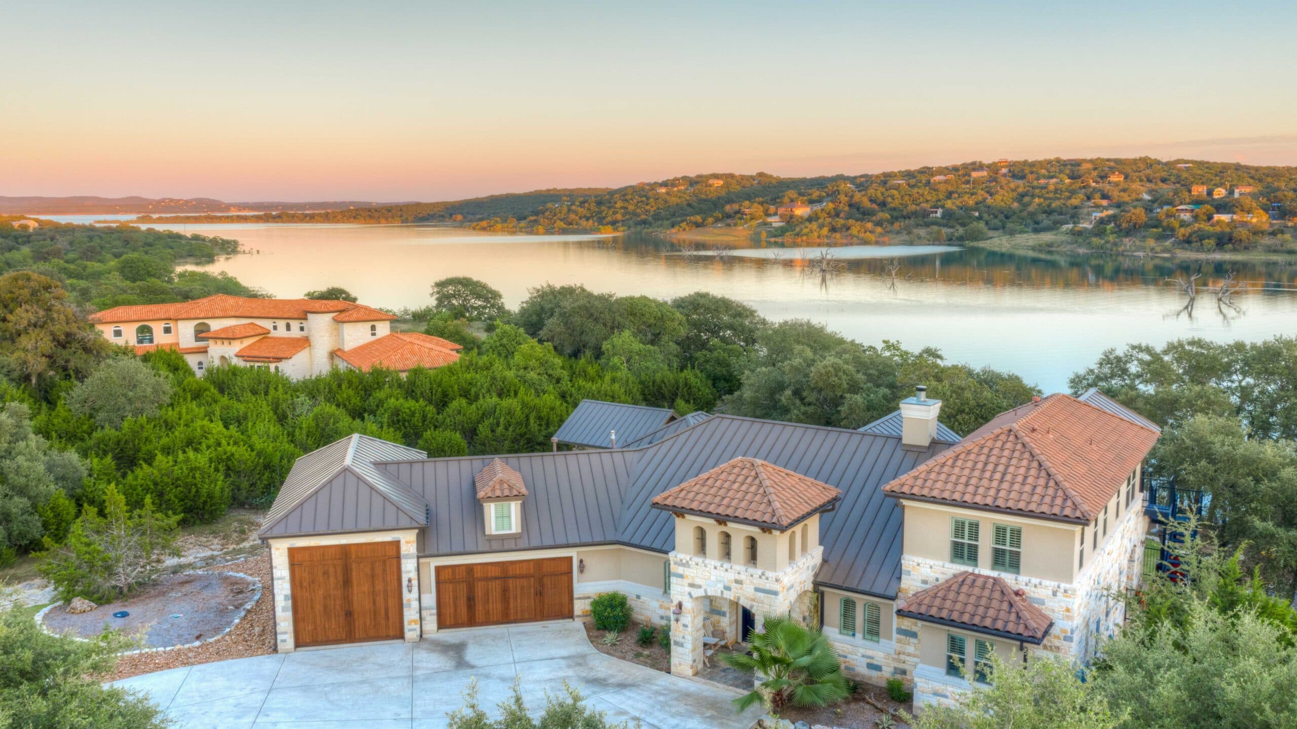 Aerial view of a residential hillside with orange-tiled roofs, a lake, and green trees at sunset
