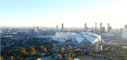 City skyline on the horizon with tall buildings, a white geometric stadium, smoke plume, and trees