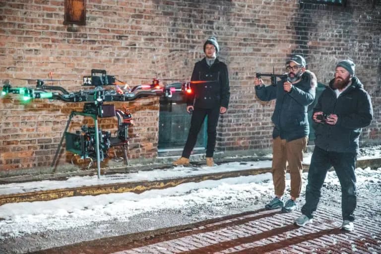 Three men in winter jackets with remote controllers near a large multi-rotor drone on a snowy brick street