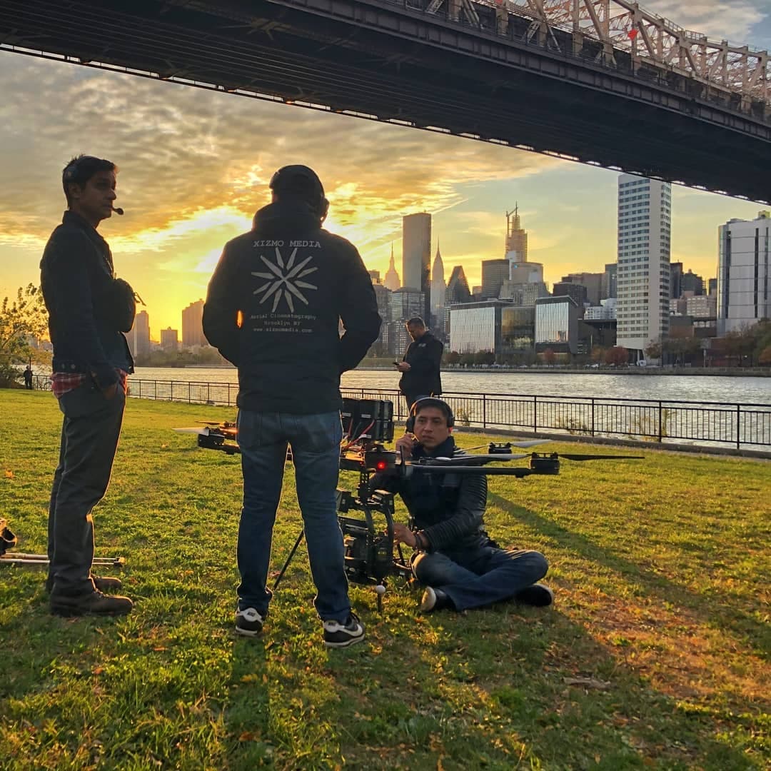 Xizmo Media crew by river at sunset, three people around a large drone and gear, skyline and bridge behind
