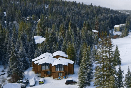 Aerial view of a snow-covered wooden cabin in a forest with several cars nearby