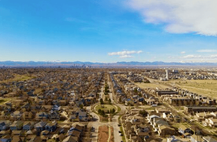 Aerial view of a grid of suburban houses with a central road, open fields and distant mountains