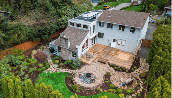 Aerial view of a two-story house with wooden deck, stone paths, circular fire pit, and landscaped backyard