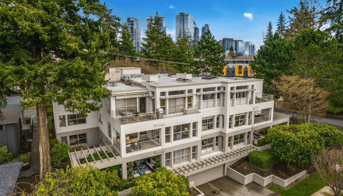 Aerial drone view of a white multistory apartment building with balconies, trees, and a city skyline