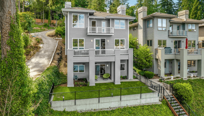 Three-story gray house with white trim, balconies, lawn, and a metal fence; hillside driveway left