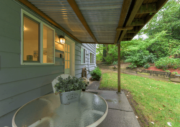 Front porch of a blue house with a glass door, windows, and a round glass table holding a potted plant; lawn and trees in background