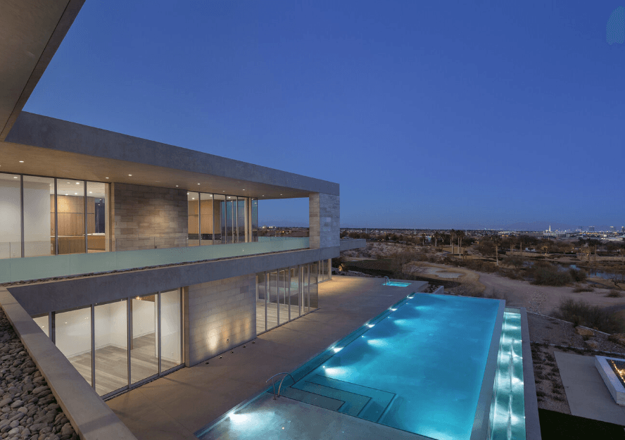 Twilight aerial view of a modern building with glass walls and a lit pool along a terrace