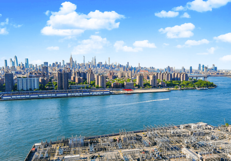 Drone view of a city skyline along a wide river with power substation equipment in the foreground