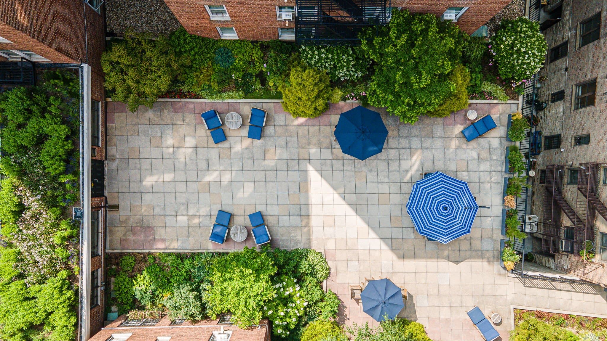Aerial view of a tiled courtyard with blue lounge chairs, round tables, and umbrellas, surrounded by greenery
