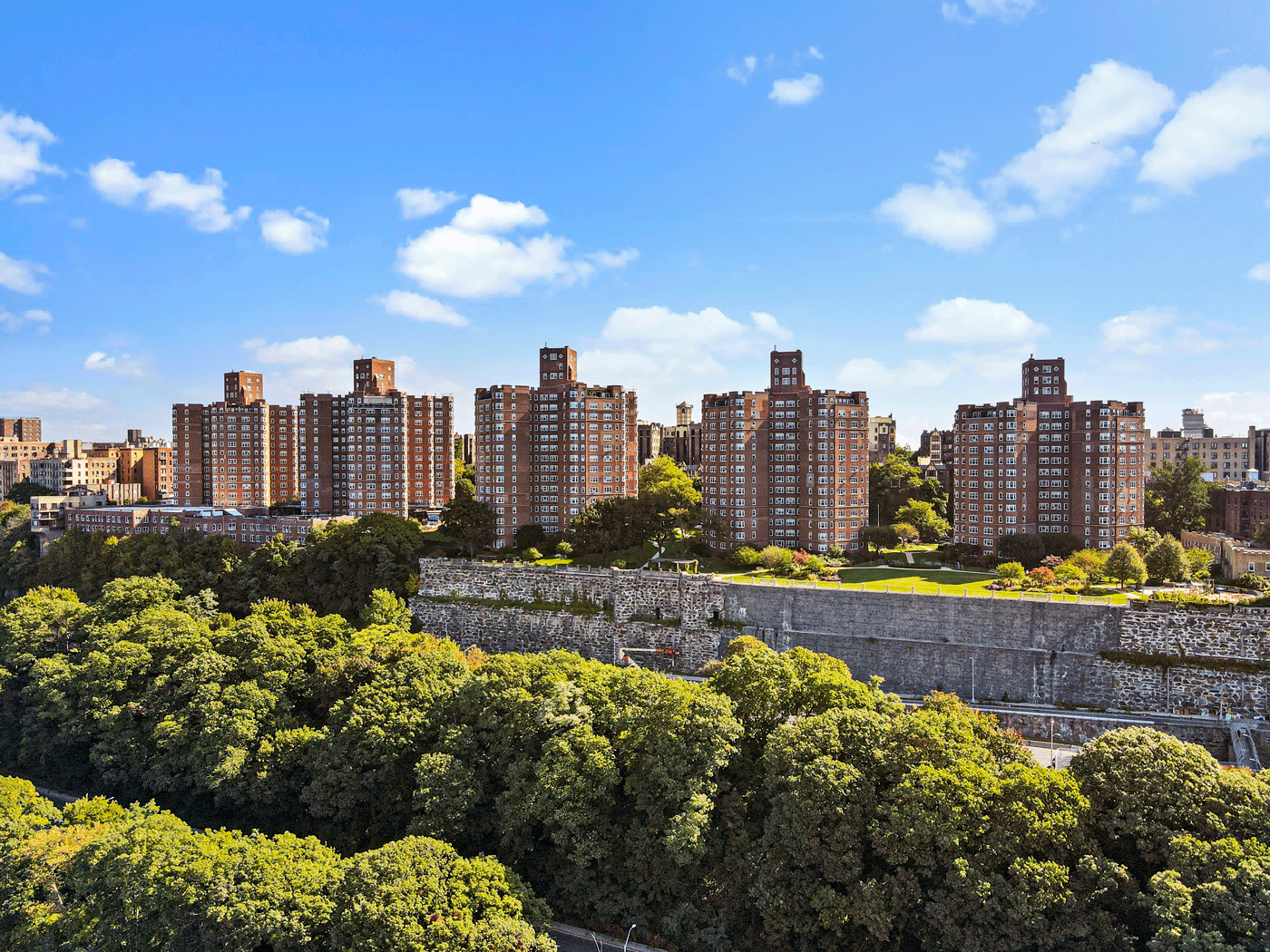 Row of tall brick apartment buildings along a green park with trees and a stone wall in foreground