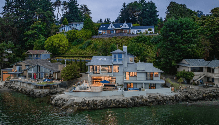 Modern waterfront home on a rocky shore with a deck and patio, surrounded by trees and neighboring houses