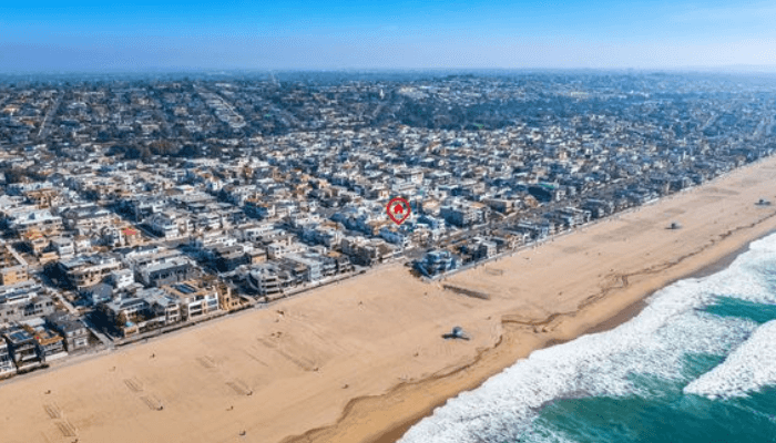 Aerial view of a beach town with a long sandy shore, waves, and row houses along the coast with a red pin