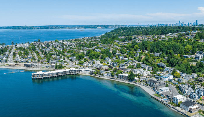 Aerial view of a coastal town with houses along the shoreline, a pier, and distant city skyline