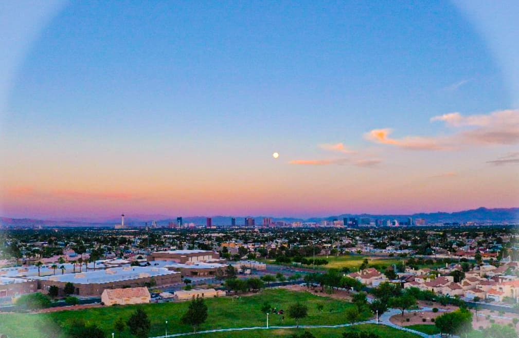 Aerial view of a suburban landscape with houses and trees, a distant city skyline at sunset, and a visible moon
