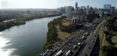Aerial view of a river on the left, with a city skyline and a busy multi-lane highway along the right