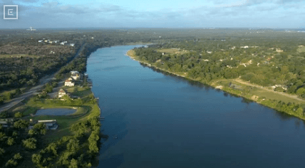 Aerial view of a winding river with green banks, houses along the shore, and fields in the distance