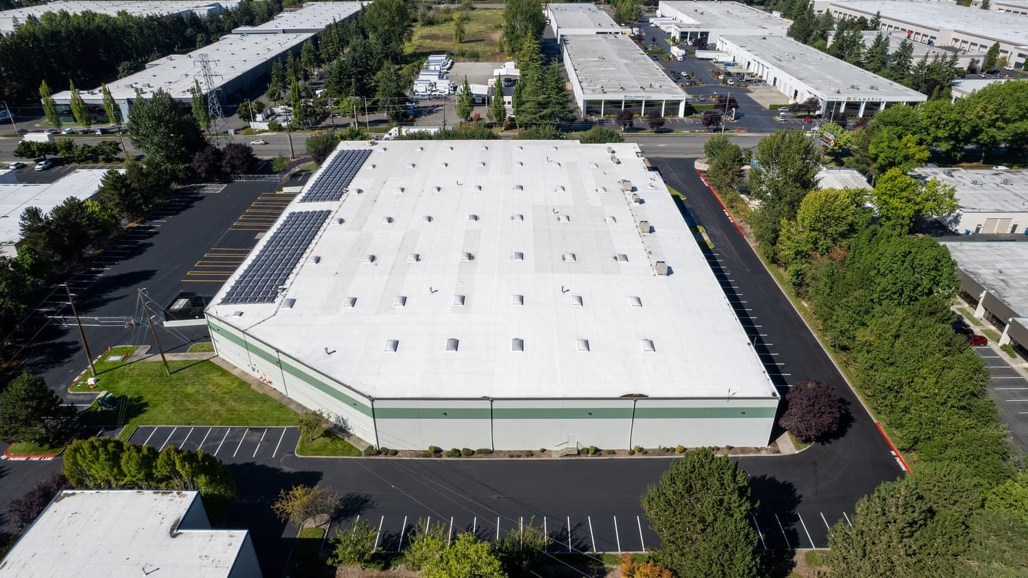 Aerial view of a white flat-roof building with skylights and solar panels, surrounded by parking and trees