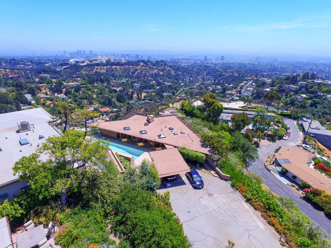 Aerial view of a hillside neighborhood with brown roofs, a house with a pool, and a distant city skyline