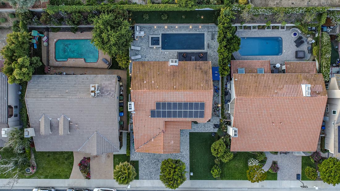 Aerial view of suburban rooftops with solar panels, backyards with pools, and manicured lawns