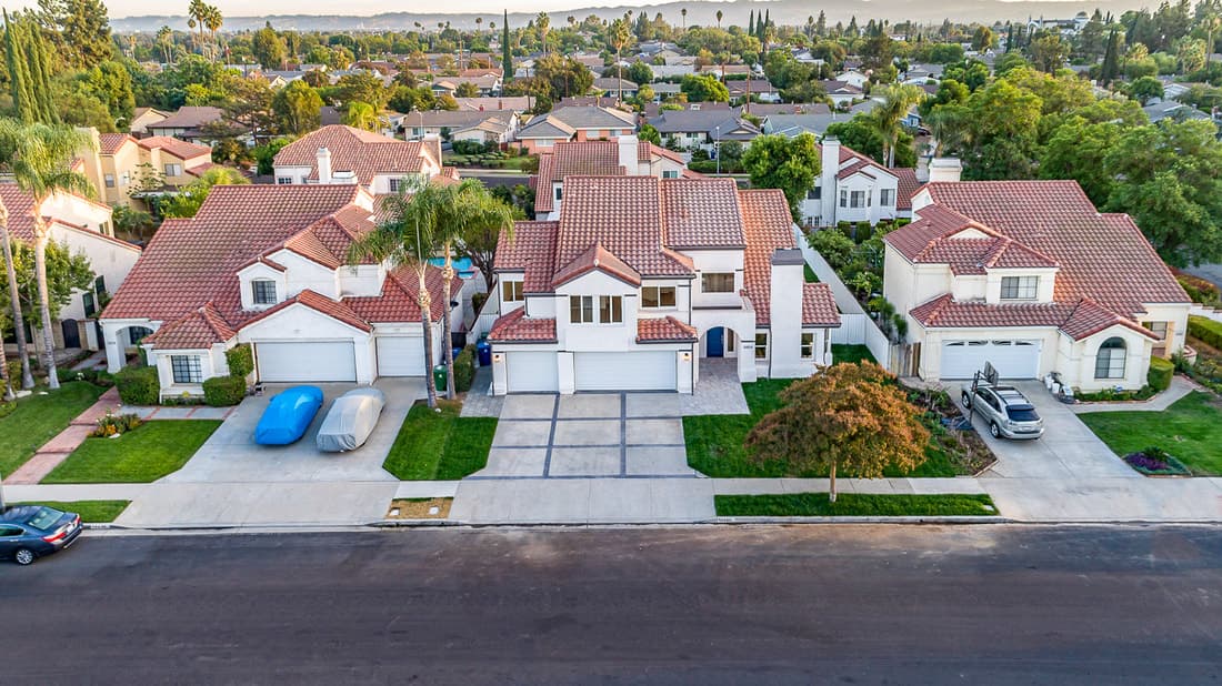 Aerial view of a suburban street with white stucco houses and red tile roofs, palm trees, and parked cars