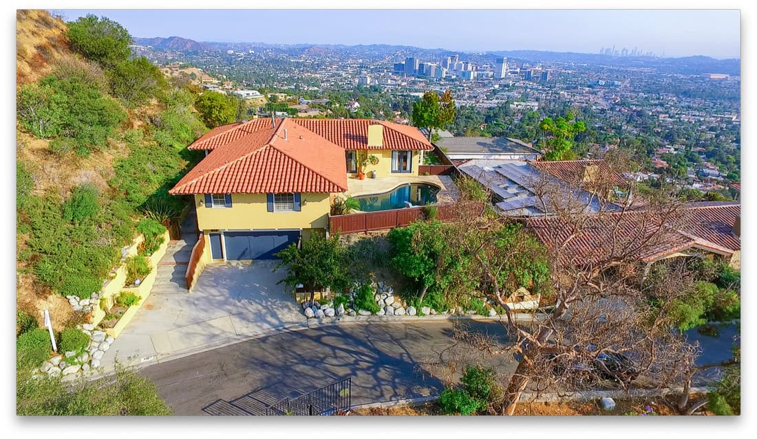 Aerial view of a yellow house with a red tile roof, curved pool, and hillside driveway, city beyond