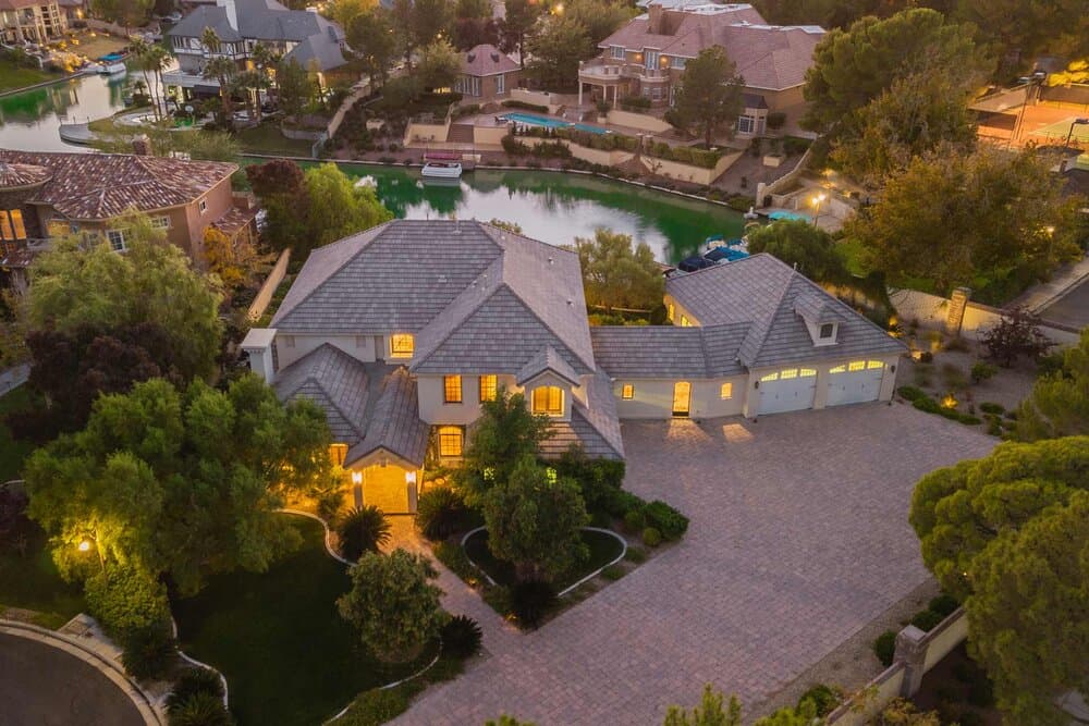 Large home with multiple gabled roofs, illuminated entry, curved stone driveway, and trees by a canal