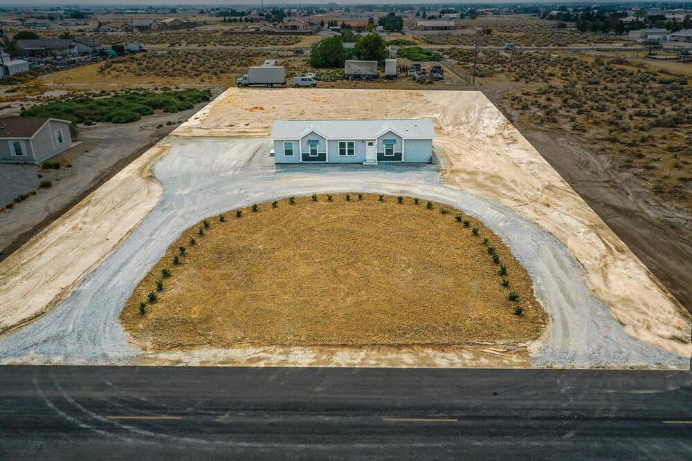 Blue single-story house with white trim, circular gravel driveway around a lawn with shrubs; desert surrounds