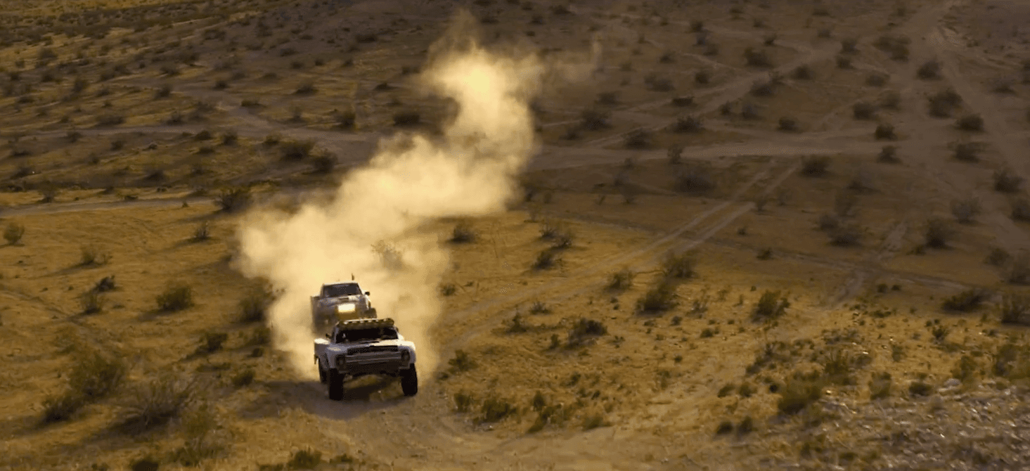Two off-road trucks drive along a dusty desert trail, raising clouds of dust