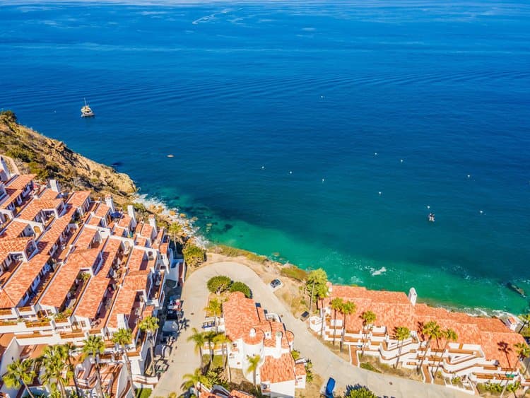 Aerial view of red-roofed houses along a rocky coastline, turquoise water, a boat, and palm trees