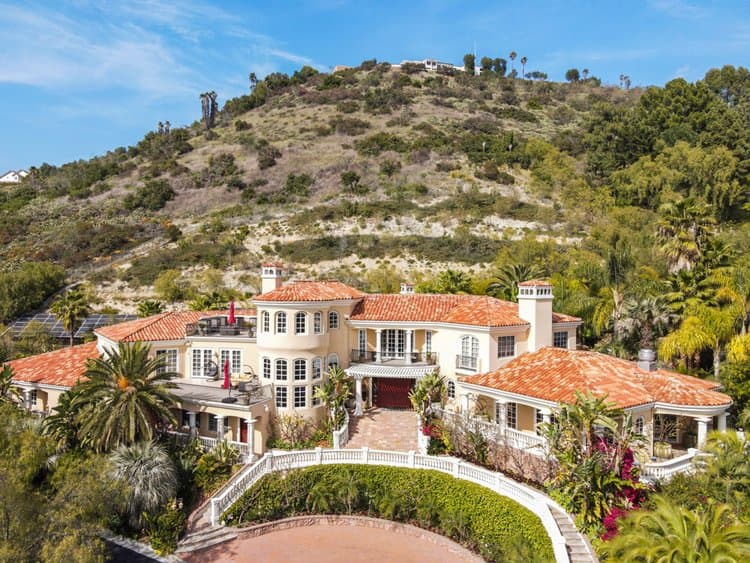 Villa with red-tiled roof on a hillside, surrounded by palm trees and landscaping, blue sky