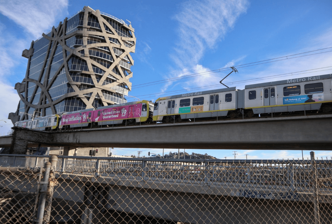 A light rail train on elevated track beside a building with crisscross concrete frame and a chain-link fence