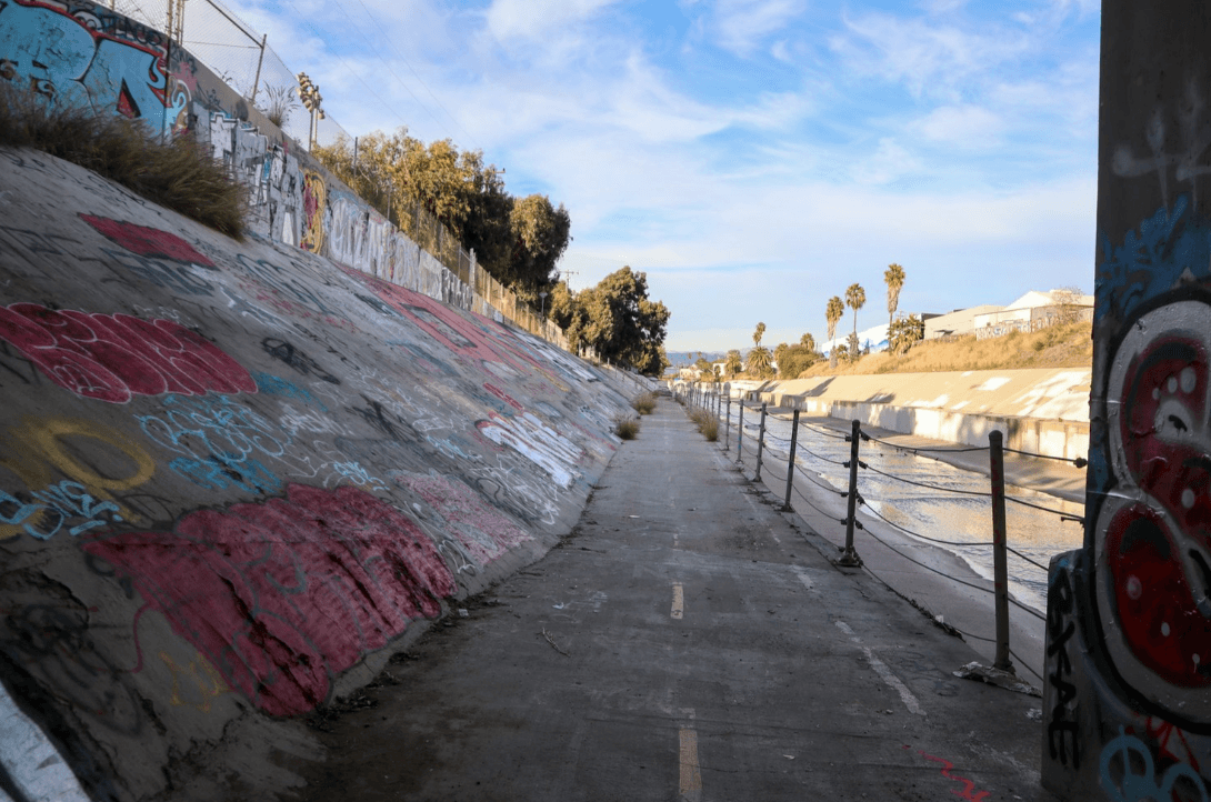 Graffiti-covered concrete canal with a narrow path and railing along the water under a blue sky