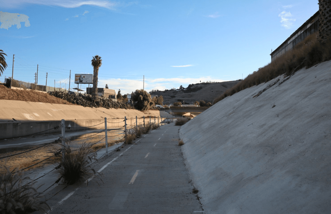 Concrete canal, pedestrian path with railing, plants along the edge, white embankment on the right, blue sky