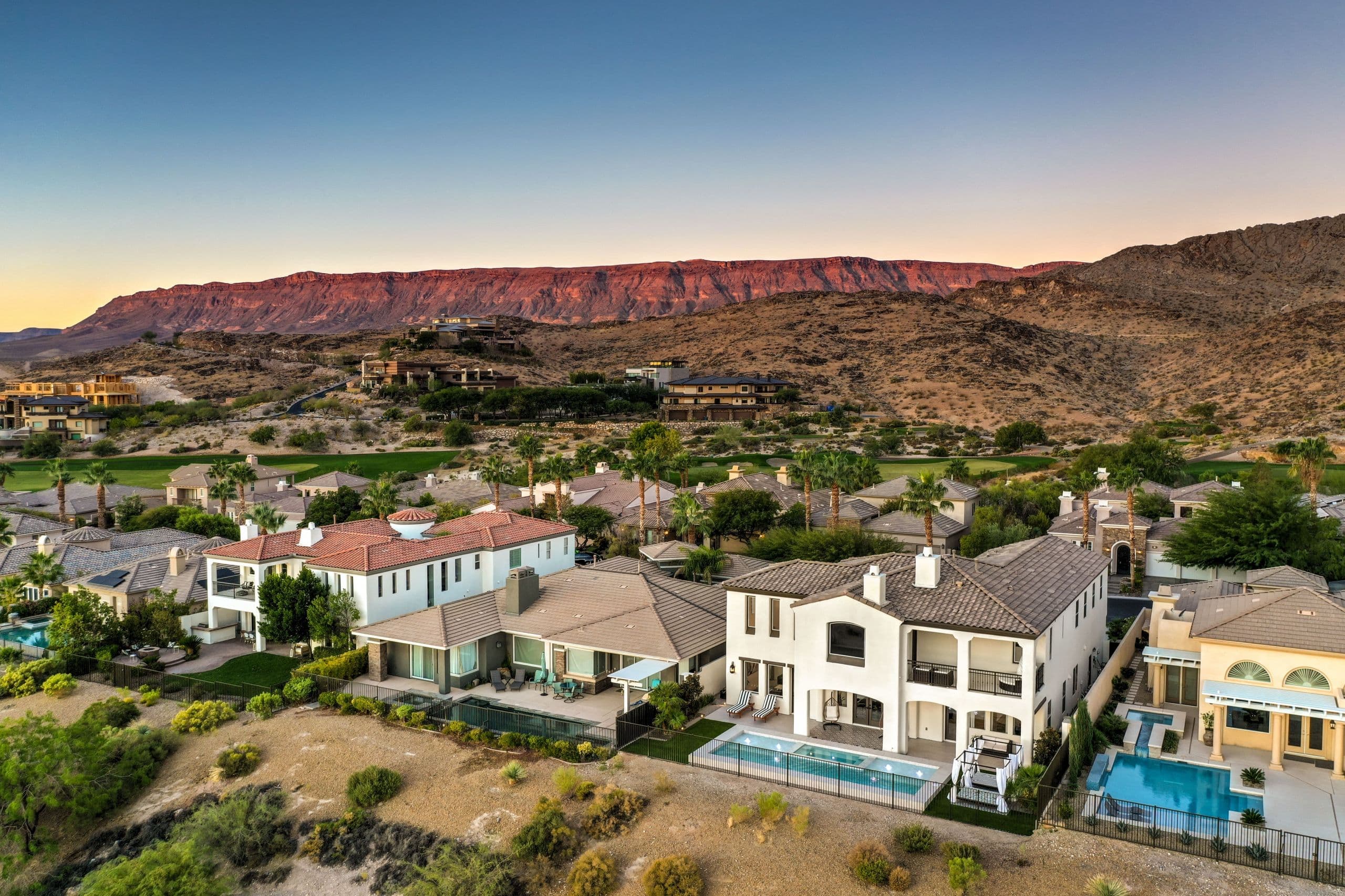 Desert suburb with tile roofs, palm trees, and pools, backdrop of red mountains at sunset