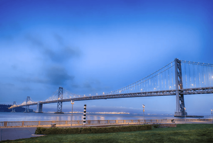 Blue hour view of a suspension bridge over water, with illuminated cables and a railing-lined grassy foreground