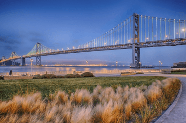 Suspension bridge at dusk over a calm river, with a curved path and tall grasses in the foreground