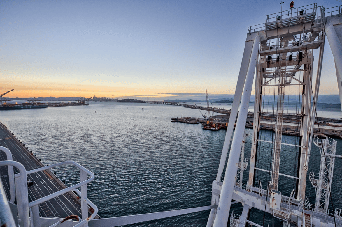 Harbor scene with a large white gantry crane on the right, calm water, docks, and distant cranes at sunset