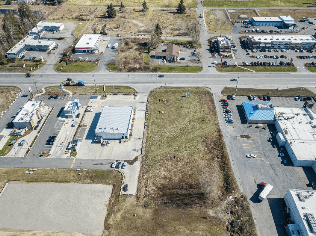 Aerial view of a commercial plaza with several buildings, parking lots, a central empty lot, and surrounding roads