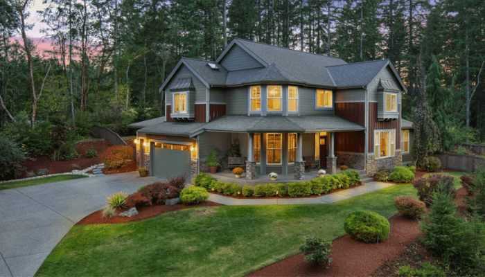 Two-story house with gray siding, stone porch, lit windows, curved walkway, and landscaped front yard at dusk