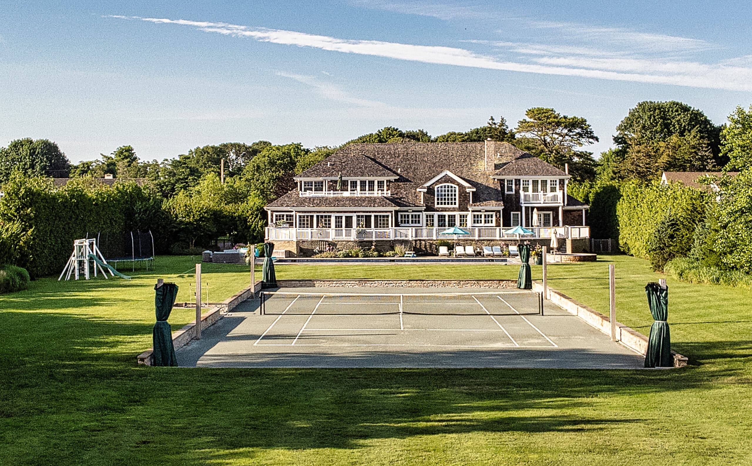 Two-story house with wraparound deck and arched window, surrounded by trees; tennis court in the foreground
